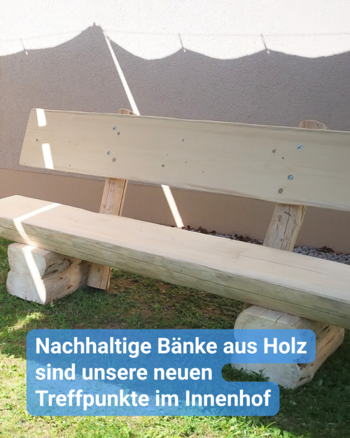 Eine Parkbank aus Holz – welche wie neu aussieht. Sie steht auf der grünen Wiese, jedoch mit dem Rücken an einer Hauswand gestellt.  Es ist ein sonniger Tag, der Sonnenschirm wirft Schatten auf die Bank. Bildbeschriftung: Nachhaltige Bänke aus Holz sind unsere neuen Treffpunkte im Innenhof