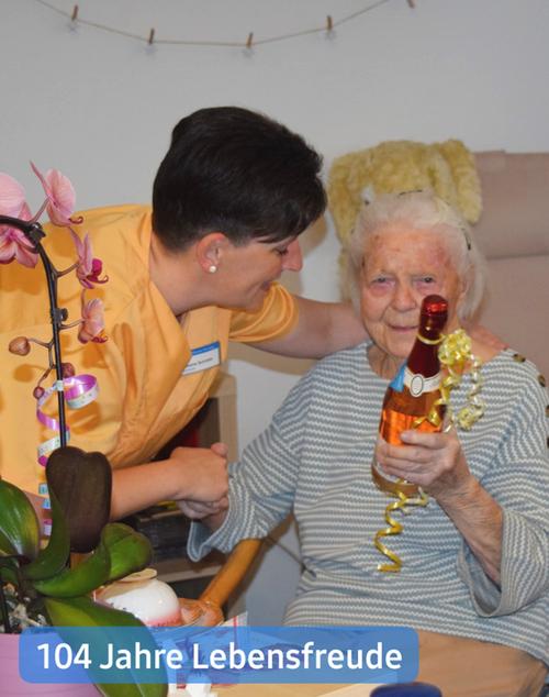 Nahaufnahme einer älteren Frau mit grauem Haar, die einen hellgrau-weiß gestreiften Pullover trägt, sitzt lächelnd da. In ihren Händen hält sie eine kleine Flasche Sekt oder Schaumwein, die mit einem gelben Band dekoriert ist.  Neben ihr sitzt eine jüngere Frau mit dunklem Haar, die ein pfirsichfarbenes, kurzärmeliges Oberteil trägt und ihren Arm liebevoll um die Schulter der älteren Dame legt. Der Gesichtsausdruck der jüngeren Frau strahlt Wärme und Fürsorge aus.  Im Hintergrund sind verschwommene Elemente zu erkennen, darunter ein Stofftier und ein Teil einer Wand. Im Vordergrund links steht ein bepflanzter Orchideenstock mit rosa und lila Blüten, der teilweise den unteren Teil der jüngeren Frau verdeckt. Am unteren Rand ist ein kleines Schild mit der Aufschrift „104 Jahre Lebensfreude“ zu sehen. Die gesamte Szenerie scheint sich in einer Wohn- oder Pflegeeinrichtung zu befinden.
