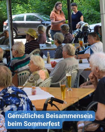Das Bild zeigt eine Gruppe von Menschen, hauptsächlich Senioren, die bei einem Sommerfest versammelt sind. Sie sitzen an Tischen unter einem Zeltdach, während im Hintergrund Bäume und ein parkendes Auto sichtbar sind. Einige Personen führen lebhafte Gespräche, während andere in unterschiedliche Richtungen blicken. Auf den Tischen stehen Gläser mit Bier und anderen Getränken, was auf eine entspannte und gesellige Atmosphäre hindeutet. Der Gesamteindruck vermittelt eine angenehme und herzliche Zusammenkunft, geprägt von Gemeinschaftsgeist und Freude unter den Teilnehmenden. Ein blaues Banner mit dem Text „Gemütliches Beisammensein beim Sommerfest” ist am unteren Rand des Bildes sichtbar.