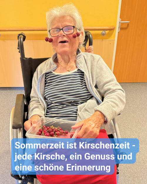 Seniorin im Rollstuhl hält stolz die Kirschernte in einer Schale auf dem Schoß fest. Sie lächelt fröhlich in die Kamera. Kirschen hängen dekorativ an ihrer Brille herab. Bildbeschriftung: Sommerzeit ist Kirschenzeit - jede Kirsche, ein Genuss und eine schöne Erinnerung